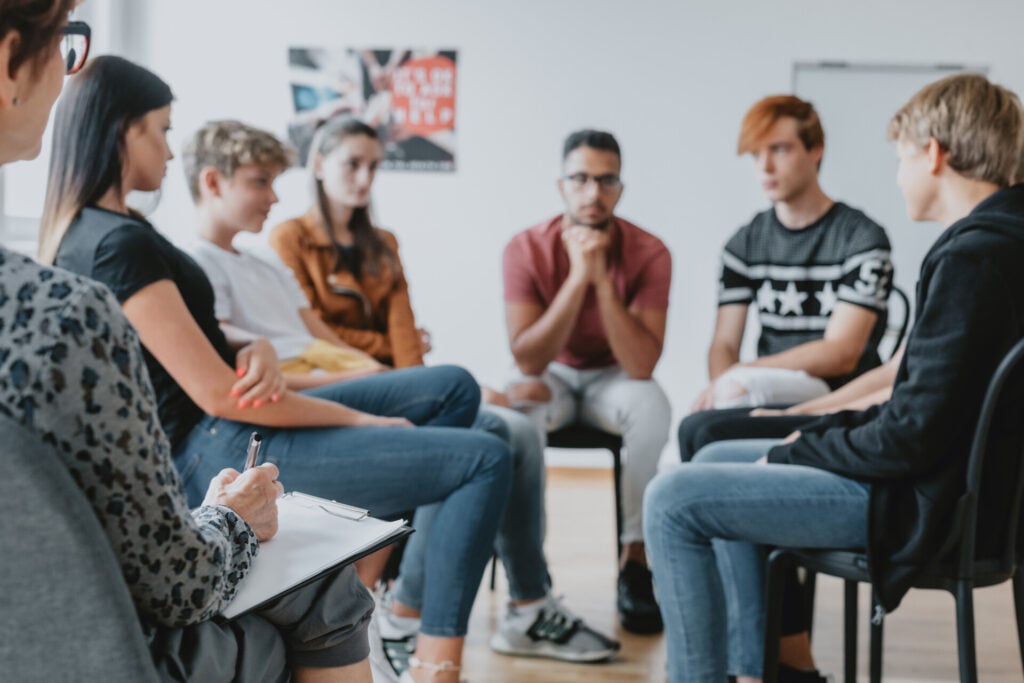 a focus group of people sat on chairs in a circle. one person is making notes on a clipboard.