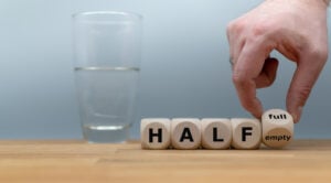 Hand turns a dice and changes the expression "half empty" to "half full" with a glass of water in the background